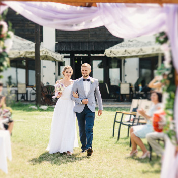 Щасливий момент відразу після обміну обітницями 💍. Молодята йдуть від весільної арки під радісні оплески гостей. Живий репортажний кадр з виїзної церемонії просто неба, що ідеально передає щирі емоції, сонячну атмосферу літнього свята та динаміку моменту. Наречена в легкій білій сукні з ніжним букетом, наречений у стильному світлому піджаку та метелику.