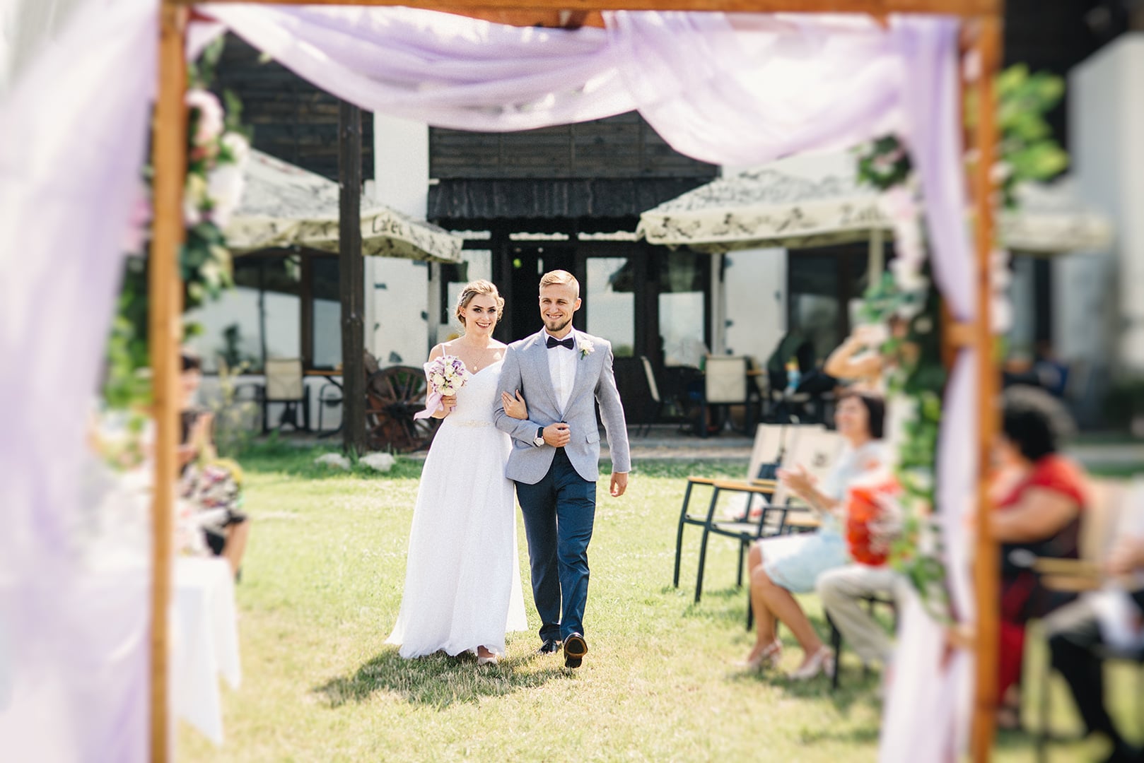 Щасливий момент відразу після обміну обітницями 💍. Молодята йдуть від весільної арки під радісні оплески гостей. Живий репортажний кадр з виїзної церемонії просто неба, що ідеально передає щирі емоції, сонячну атмосферу літнього свята та динаміку моменту. Наречена в легкій білій сукні з ніжним букетом, наречений у стильному світлому піджаку та метелику., фото - Фотограф Дмитро Покідін