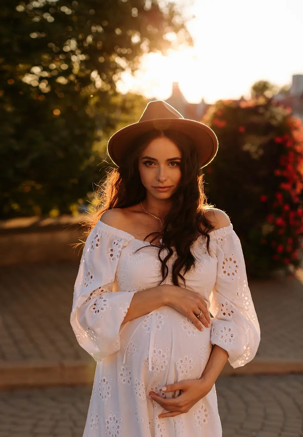 Вулична фотосесія вагітності в білій сукні і капелюху в парку на заході сонця