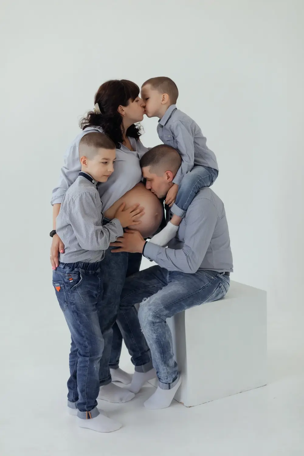 Велика сімейна фотосесія вагітності з дітьми у синіх джинсах та світлих сорочках на білому фоні