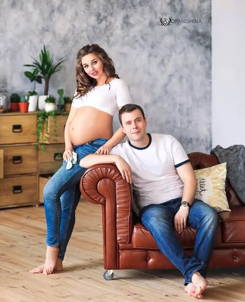 Затишна сімейна фотосесія вагітності де жінка з відкритим животом та чоловік позують сидячи на шкіряному дивані